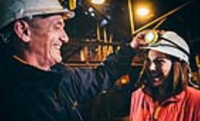 a miner in a hard hat places a white helmet on a lady wearing an orange coat, then switches on the map attached to her helmet